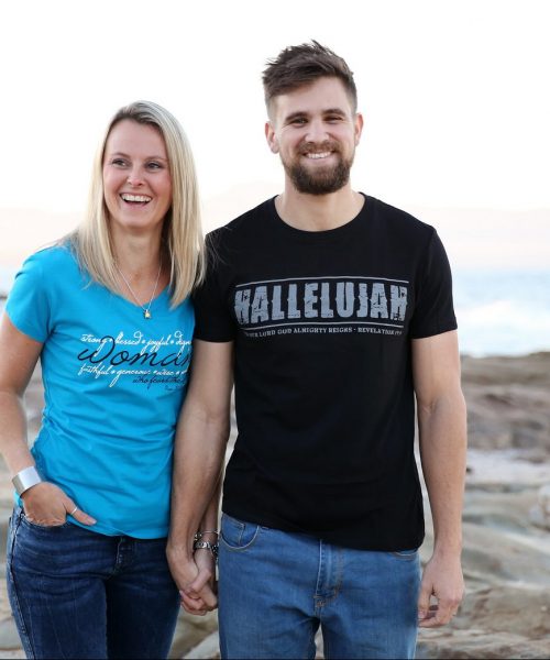 Man on beach wearing black Christian T shirt with grey words "Hallelujah for our Lord God Almighty reigns" printed on it by In The Gap Clothing